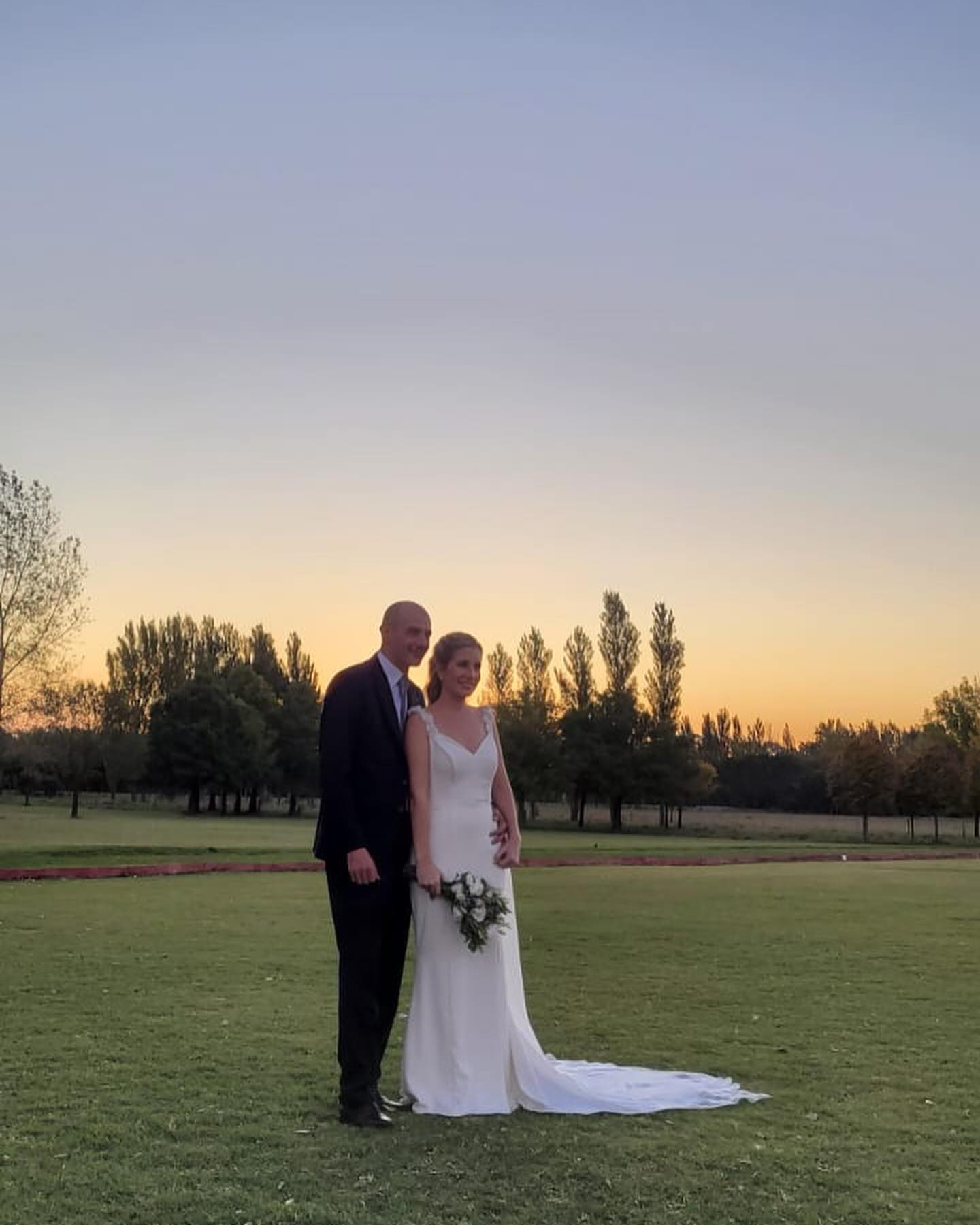 A bride and a groom on their wedding day celebration. They are standing on a field with freshly cut grass, the sunset behind them creating an peach and light blue sky. In the distance, the woods create a beautiful landscape.